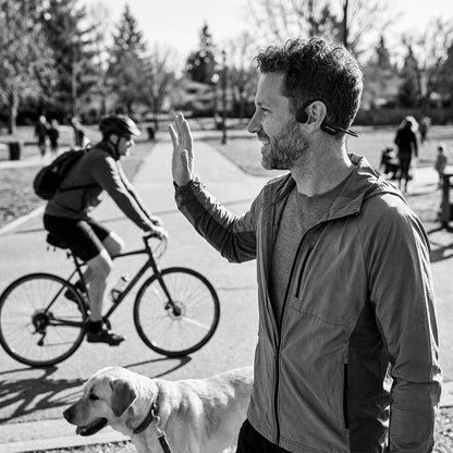 A man waves while wearing OPN Sound Osso bone conduction headphones in a park with his dog.